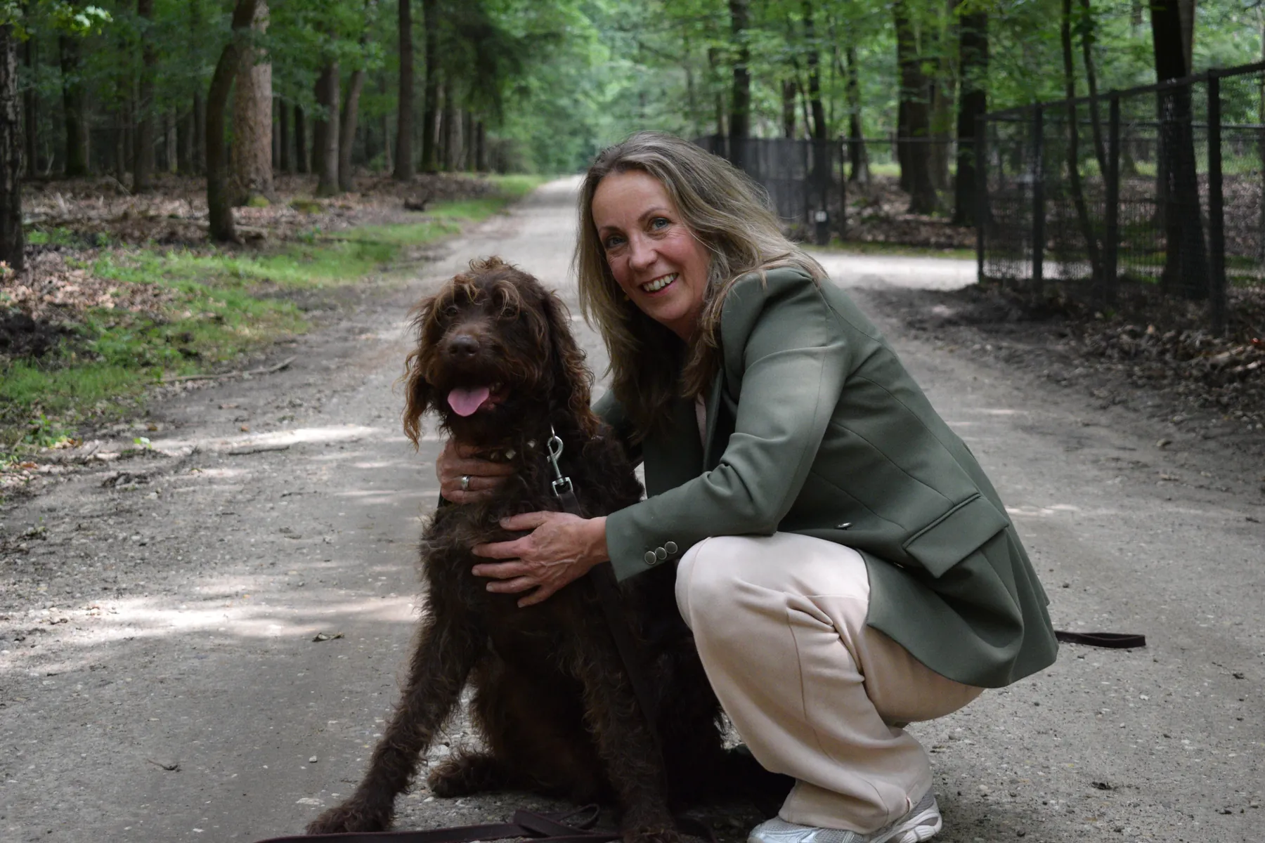 Marjon zit lachend op een bospad, met haar arm om een bruine hond