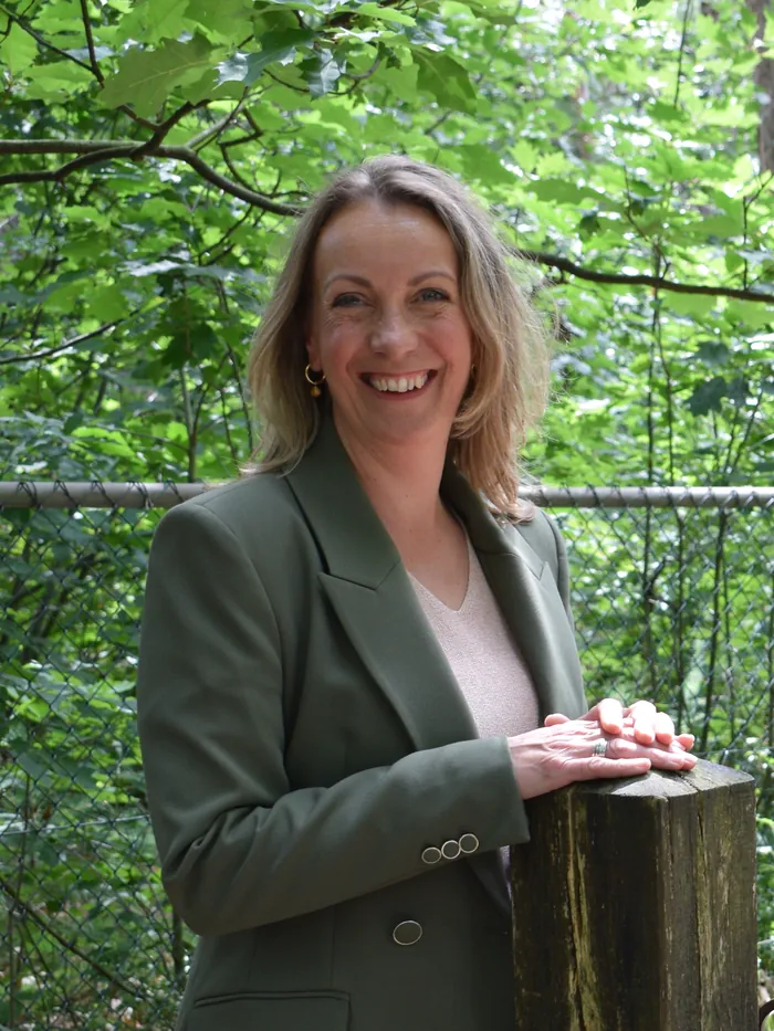Marjon Brinks kijkt je lachend aan in een groen colbert, met haar handen op een houten paal aan de rand van het bos