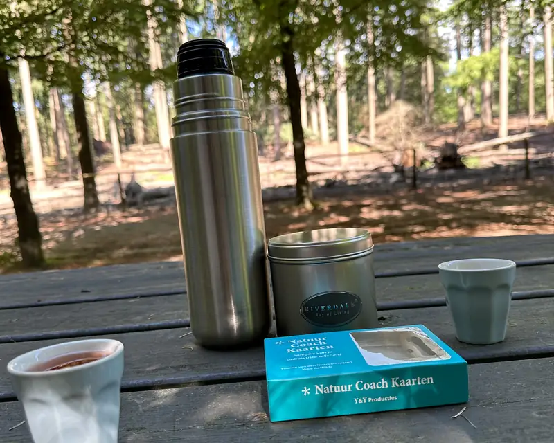 Een thermosfles, bekers en een doos Natuur Coach Kaarten op een picknicktafel in het bos