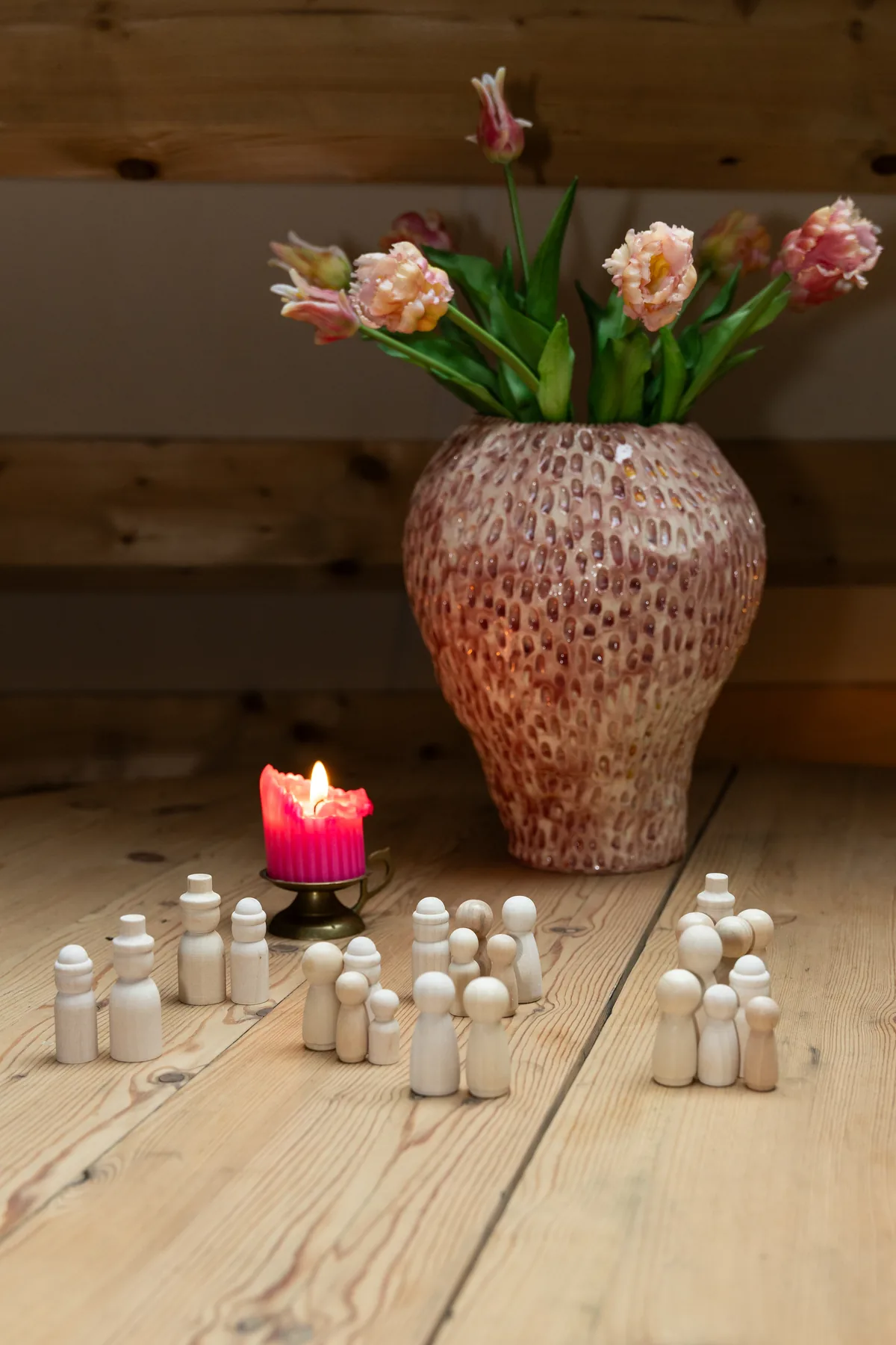 Houten poppetjes in kleine groepjes op een houten vloer, met een brandende kaars en een vaas tulpen, zoals gebruikt bij een opstelling