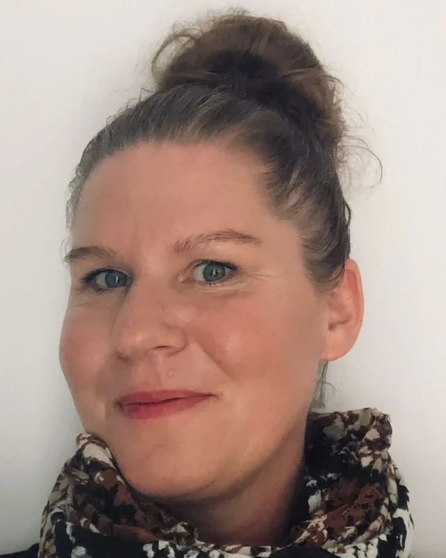Portret van Wendy Koers, glimlachend voor een lichte muur, met een sjaal om